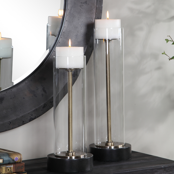 candle picks Uttermost Candleholder This Set Of Two Candleholders Feature An Aged Brass Stand Surrounded By A Clear Glass Hurricane And Finished With A Thick Bronze Foot. Two 4"x 3" Distressed Off-white Candles Included. Sizes: S- 7x16x7, L- 7x20x7
